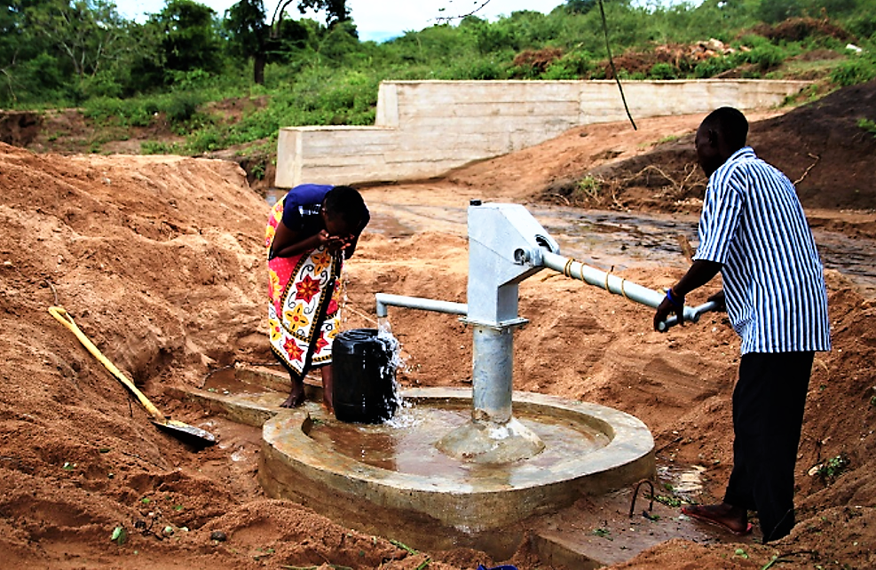 Two local people fetching water using a hand pump from a closed well near a sand dam